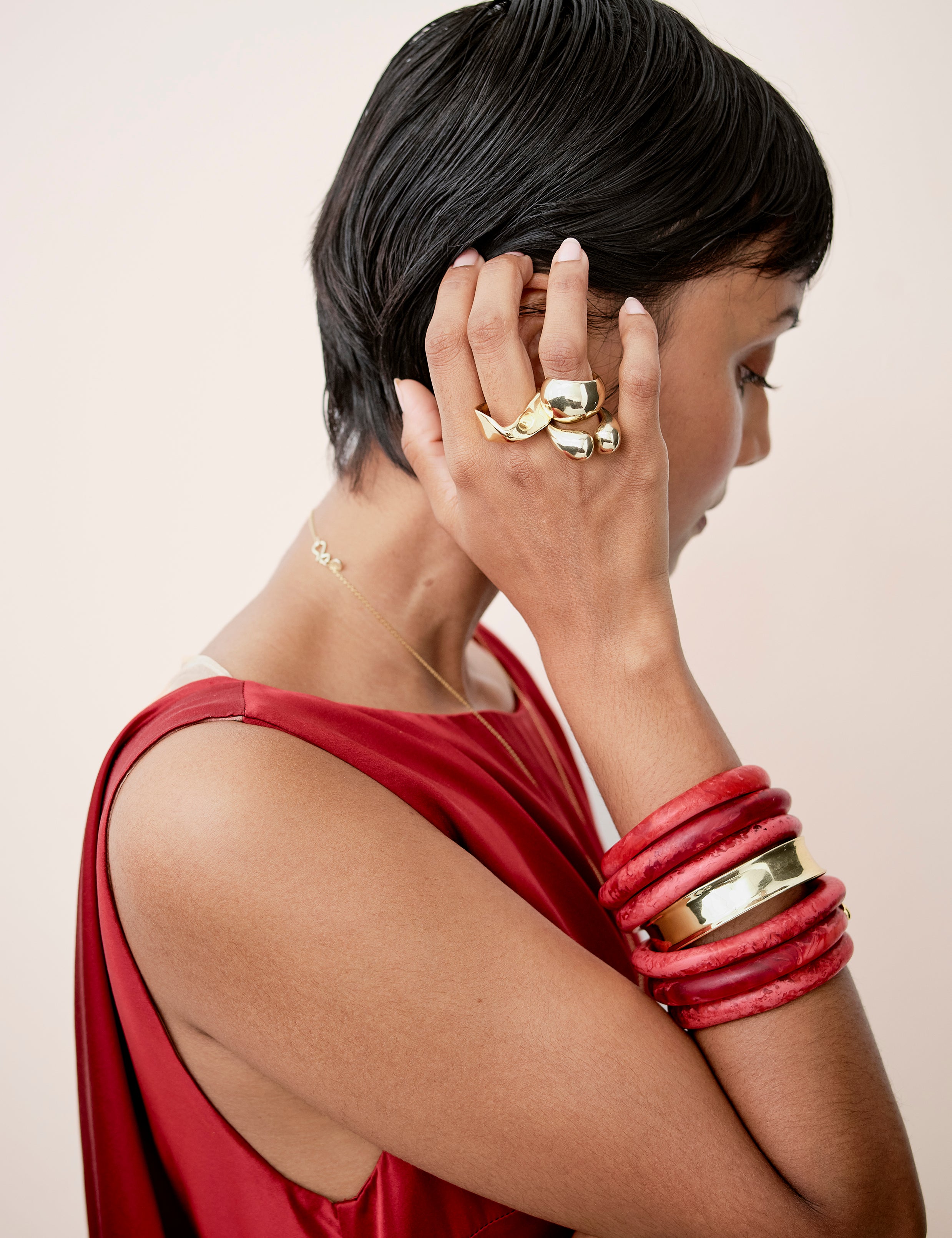 Model wearing a stack of deep red resin bangles with a single brass bangle. Accompanied by a stack of brass rings held up against ear.