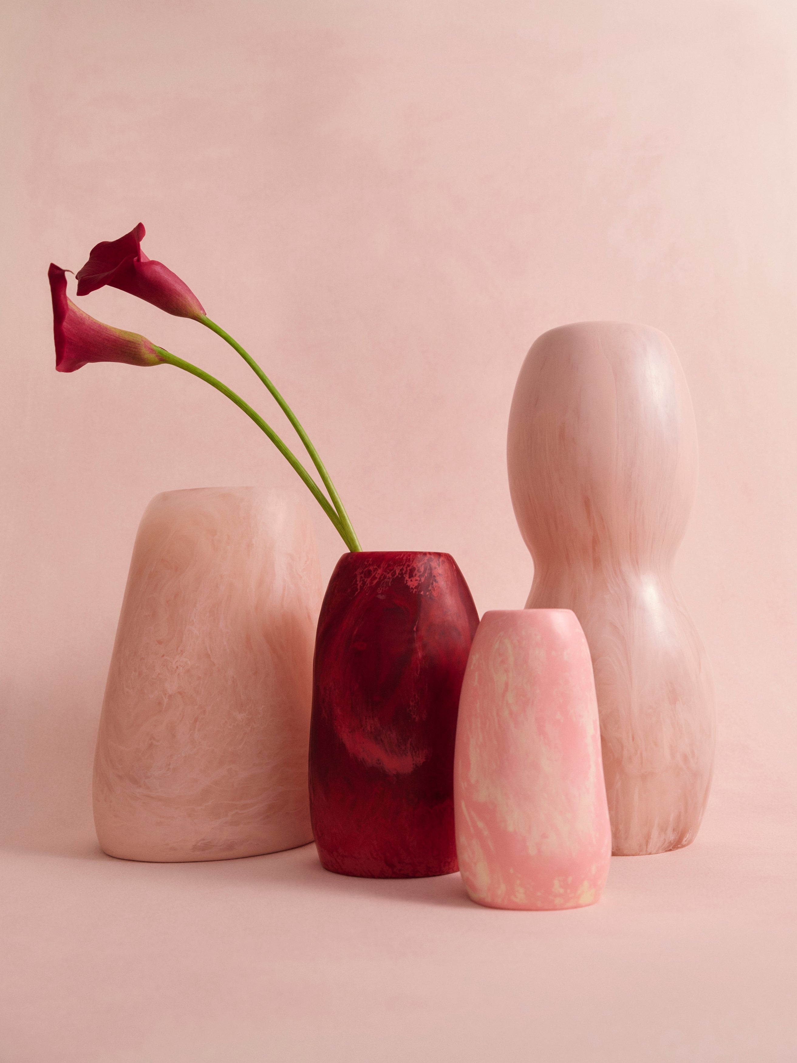 Set of four pink resin vases in a variety of organic shapes on a pink background with two floral stems